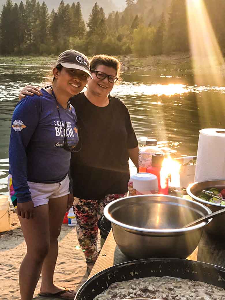 Two women smiling while standing near a serving table at dinner during a Main Salmon River whitewater rafting vacation in Idaho.